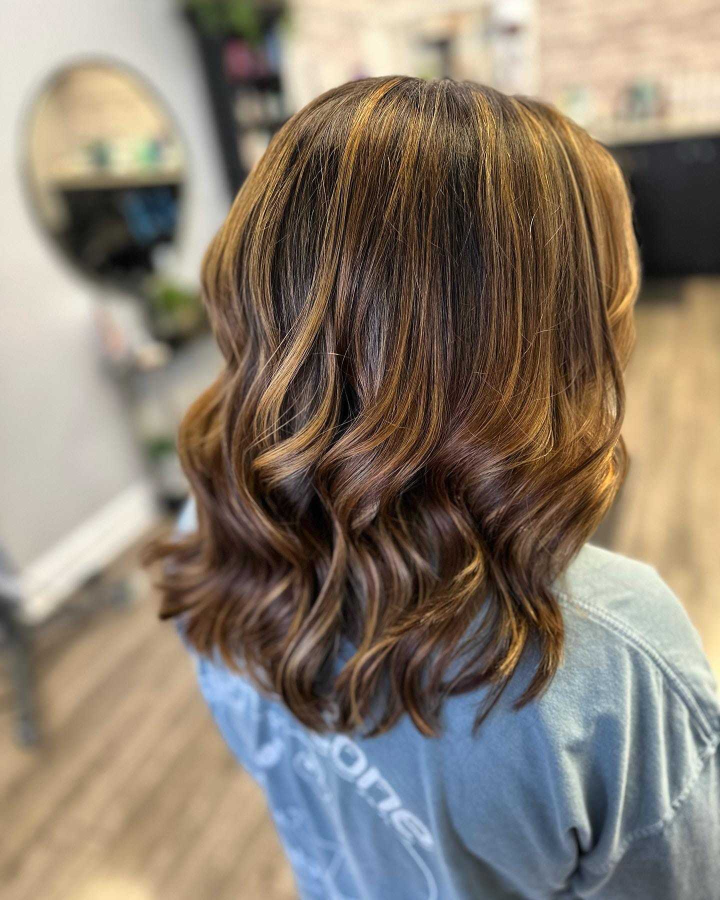 Woman with wavy, highlighted hair in a salon, facing away from the camera.