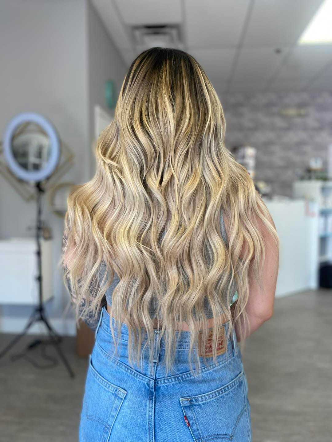 Woman with long, wavy blonde hair wearing jeans, standing in a modern salon.